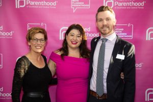 Three people stand smiling in front of a pink Planned Parenthood banner.