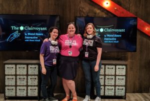 Three people wearing t-shirts with "Strong Female Character" stand in front of two displays showing "The Clairvoyant by Weird Sisters Interactive.