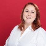 A person with red hair smiles, wearing a white shirt against a red background.