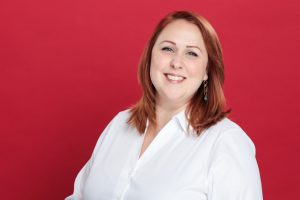 A person with red hair smiles, wearing a white shirt against a red background.