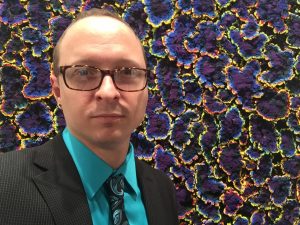 A person wearing glasses and a suit with a teal shirt and patterned tie stands in front of a vibrant, abstract background.