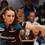 A woman bowling in a competition, wearing a black and blue outfit. Spectators are seated in the background.