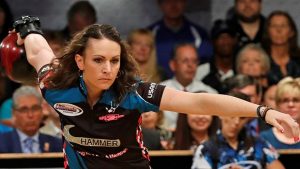 A woman bowling in a competition, wearing a black and blue outfit. Spectators are seated in the background.