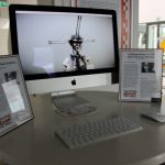 A computer on a round table displays a digital artwork. Two informational stands flank the screen, and a keyboard and mouse are on the table.