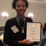 Person holding a framed award in a conference room, wearing a necklace and dark sweater.