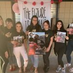 A group of seven people stand in front of a sign reading "The Future Is Female," holding magazines and smiling. Some wear matching "Fem Magic" shirts.