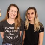 Two women stand in front of a gray wall. One wears a "Women change the world" t-shirt, and the other wears a black top. A sign on the wall reads "100K The Gallery.