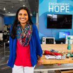 Woman smiling, standing by a table with prosthetic limbs and parts, in an office with "Engineering Hope" on the wall.