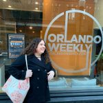 A person with curly hair and a tote bag stands in front of a glass door proudly featuring the iconic "Orlando Weekly" logo.