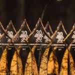 A row of transparent 30 Under 30 award trophies with engraved text, lined up on a dark surface.