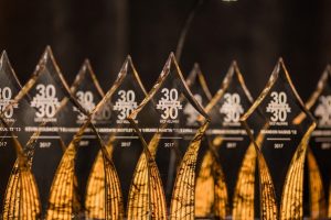 A row of transparent 30 Under 30 award trophies with engraved text, lined up on a dark surface.