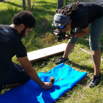 A man kneels on the grass beside a blue mat holding a pine cone, while a woman photographs it with a camera.