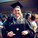 A person in a graduation cap and gown holds a diploma folder, standing outdoors among a crowd.