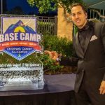 A man in formal attire poses next to an ice sculpture with the "BASE Camp Children's Cancer Foundation" logo.