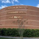 Brick building with the sign "Nicholson School of Communication and Media" and shrubs in front. A person walks on the sidewalk.