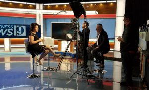 A woman in a polka dot dress is being interviewed in a TV studio. Three men operate cameras and equipment around her. A large screen displays "News 9" in the background.