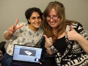 Two people smiling and gesturing thumbs up and peace sign, sitting in front of a laptop displaying a design software.