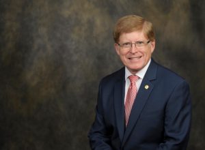 A middle-aged man with glasses, wearing a dark suit and red tie, seated against a dark mottled background, smiling at the camera.