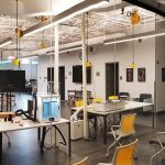 A modern laboratory with workstations, chairs, overhead lighting, and various equipment. Large screens and machinery are visible, and the space is clean and organized.