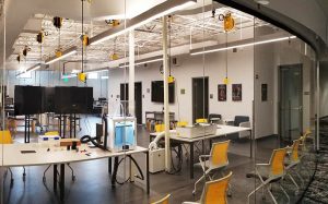 A modern laboratory with workstations, chairs, overhead lighting, and various equipment. Large screens and machinery are visible, and the space is clean and organized.