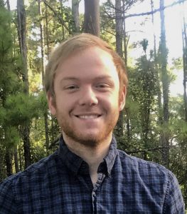 A person with light hair and a beard is smiling in front of a forest backdrop, wearing a checkered shirt.