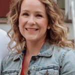 A person with curly hair smiles, wearing a denim jacket over a red top.