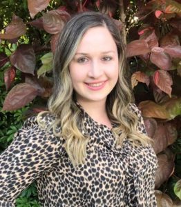 A person with long wavy hair smiles in front of leafy plants, wearing a leopard print blouse.