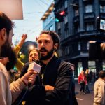 A journalist interviews a man on a busy city street. Camera equipment is visible, and pedestrians are in the background.