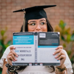 Graduate in a cap and gown holds a book in front of their face, standing against a blurred brick wall background.