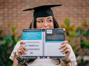 Graduate in a cap and gown holds a book in front of their face, standing against a blurred brick wall background.