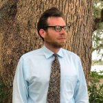 Man in glasses wearing a light blue shirt and patterned tie stands in front of a large tree trunk.