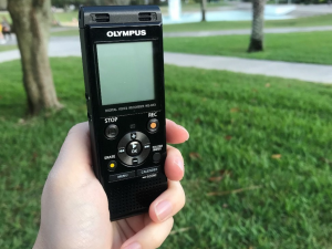 A hand holding an Olympus digital voice recorder in a park, with grass and trees in the background.