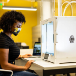 Person wearing a face mask operates a 3D printer in a modern workspace with yellow accents.