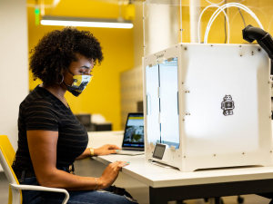 Person wearing a face mask operates a 3D printer in a modern workspace with yellow accents.