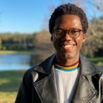 A person wearing glasses and a leather jacket stands smiling outdoors near a pond on a sunny day.