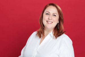 A person with red hair smiling, wearing a white shirt and earrings, against a plain red background.