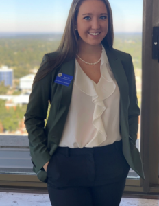 A woman in a business outfit, wearing a name badge and standing in front of a window with a cityscape in the background.