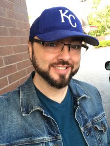 A person wearing glasses, a blue "KC" cap, and a denim jacket smiles outdoors near a brick wall.