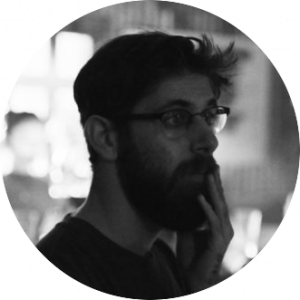 Black and white profile photo of a person with glasses and a beard, looking contemplative, with one hand touching their face. Blurred background.