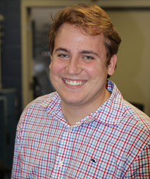 A person with brown hair smiling, wearing a checkered shirt, stands indoors.