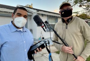 Two people wearing masks hold audio and video equipment outside a house during the day.