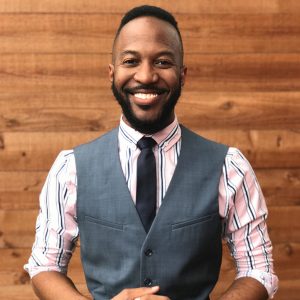 A person smiling, wearing a striped shirt and gray vest with a tie, standing in front of a wooden wall.