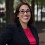 A woman with long brown hair and glasses smiles, wearing a black blazer and red top, standing outdoors.