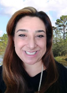 A person with long hair and a necklace smiles with a background of trees and a body of water under a blue sky.