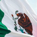 A large Mexican flag waves in the wind, featuring green, white, and red vertical stripes with an eagle on a cactus holding a snake in the center.