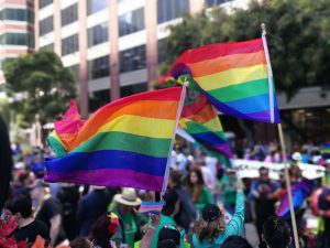 People hold rainbow flags at an outdoor event, surrounded by a crowd near tall buildings and trees.