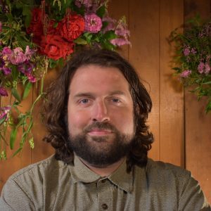 A person with long hair and a beard is standing in front of a wooden wall with colorful flowers hanging above.