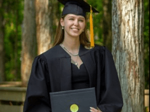 A graduate in cap and gown holds a diploma outdoors with trees in the background.