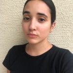 Person with dark hair tied back, wearing a black shirt, standing against a textured wall, looking directly at the camera.