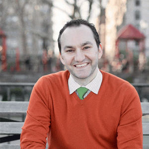 Person sitting on a bench, smiling. They wear a red sweater over a white shirt with a green tie. Blurred background shows an outdoor setting with trees and structures.